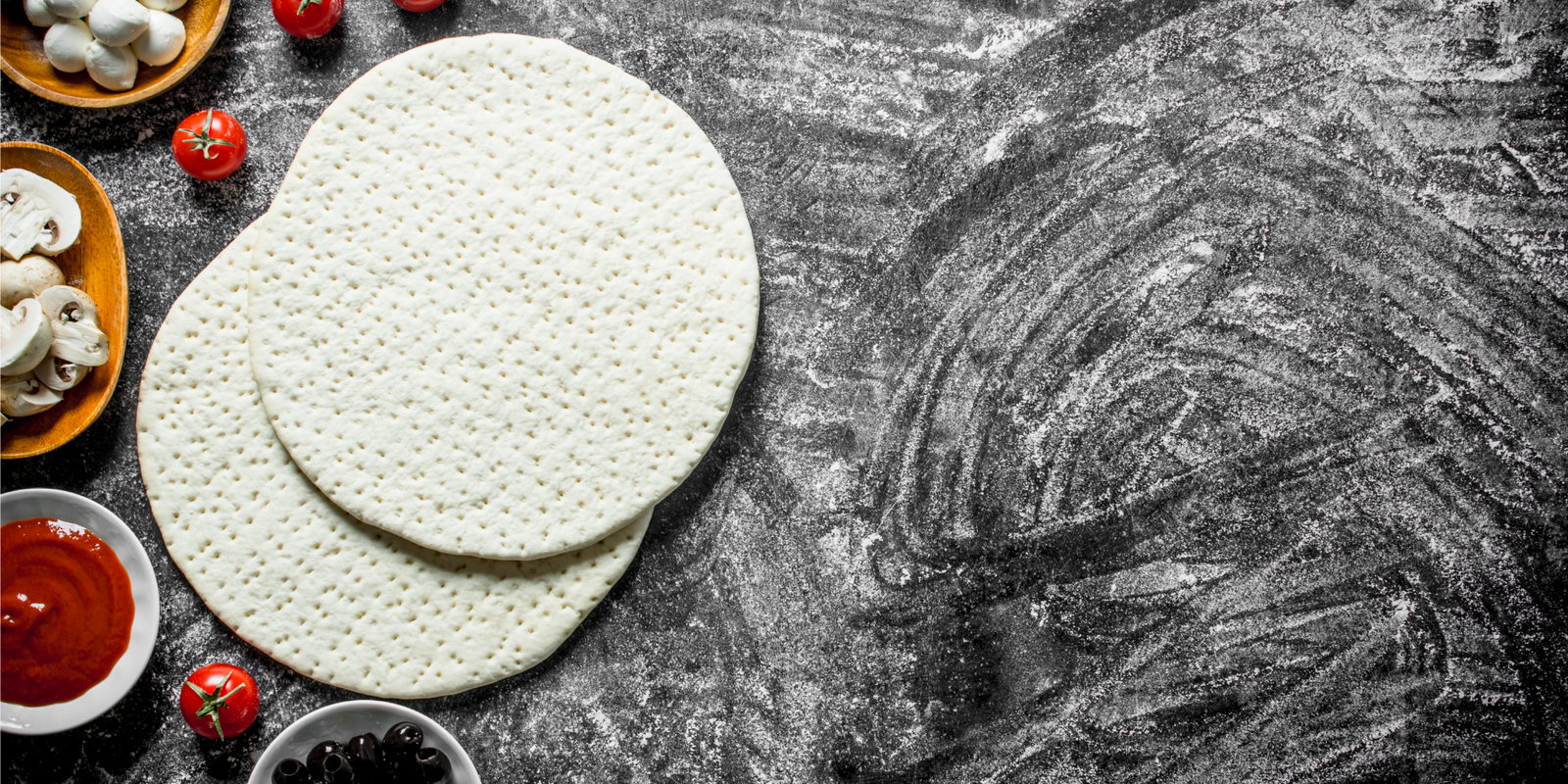 Raw pizza crust on a floured black countertop. Toppings for pizza making and tomato sauce