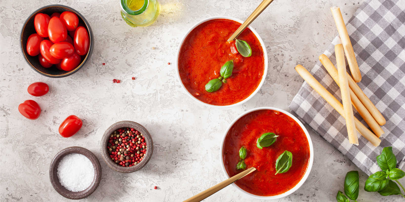 Two bowls of tomato and bell pepper soup and breadsticks on stone countertop.