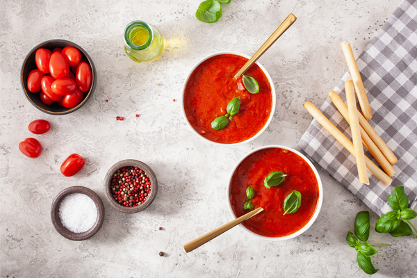 Two bowls of tomato and bell pepper soup and breadsticks on stone countertop.