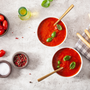 Two bowls of tomato and bell pepper soup and breadsticks on stone countertop.