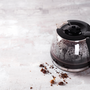 Coffee maker pot filling with coffee beans on a light stone background.