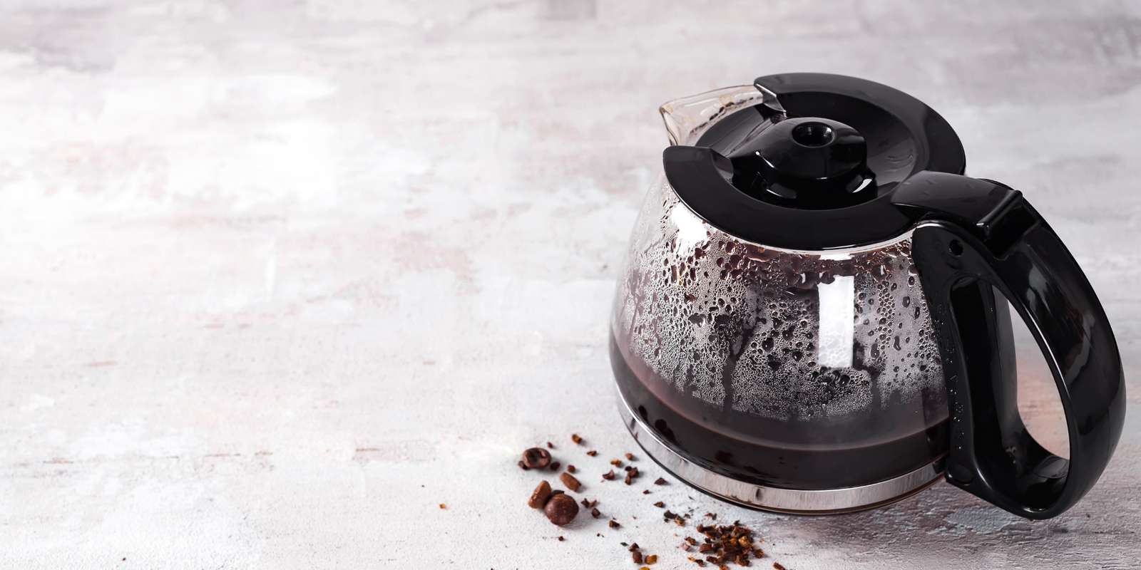 Coffee maker pot filling with coffee beans on a light stone background.