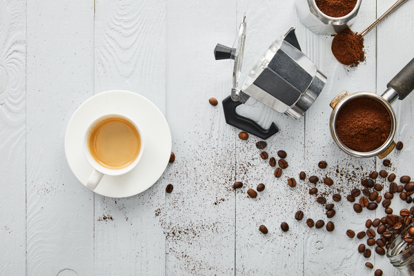 Top view of cup of coffee on saucer near geyser coffee maker, portafilter and spoon on white wooden