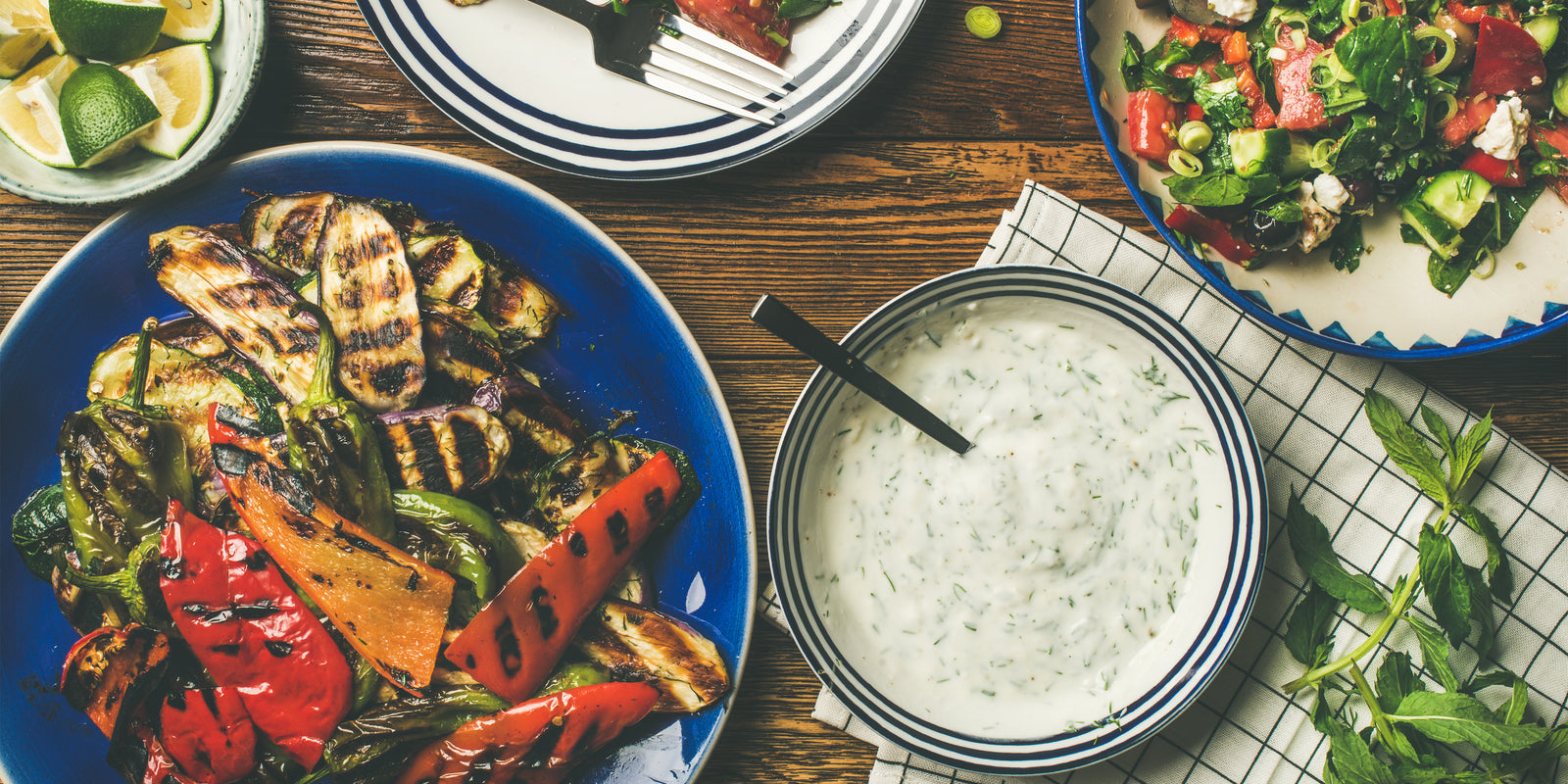 Healthy dinner table setting. Green salad, grilled vegetables with yogurt and garlic sauce, lime slices water over wooden background, top view, horizontal composition. Vegan food.