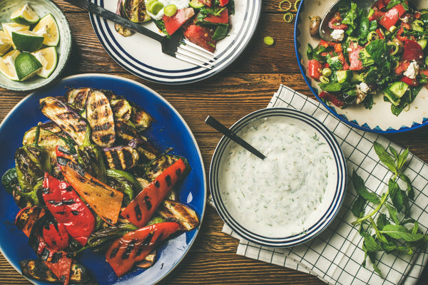 Healthy dinner table setting. Green salad, grilled vegetables with yogurt and garlic sauce, lime slices water over wooden background, top view, horizontal composition. Vegan food.
